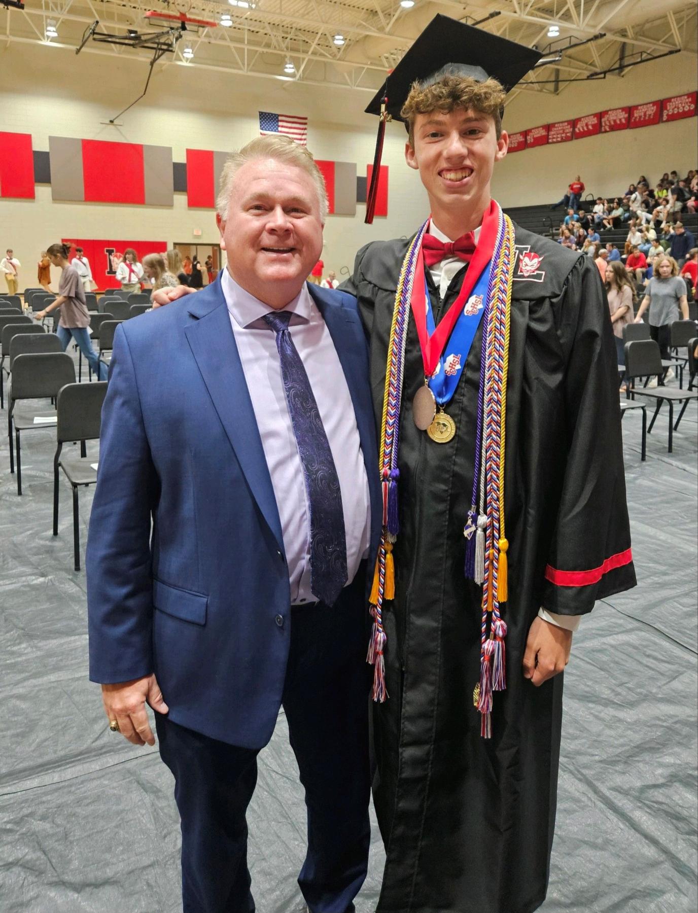 Roy Costner (left) dressed in business attire stands beside scholarship winner Isaac Esuary, who wears a graduation cap and gown. Both smile into the camera. A high school gym is in the background.