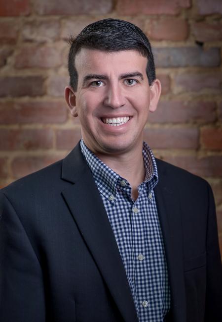 Man in a blue suit jacket with a blue button down shirt in front of a brick background