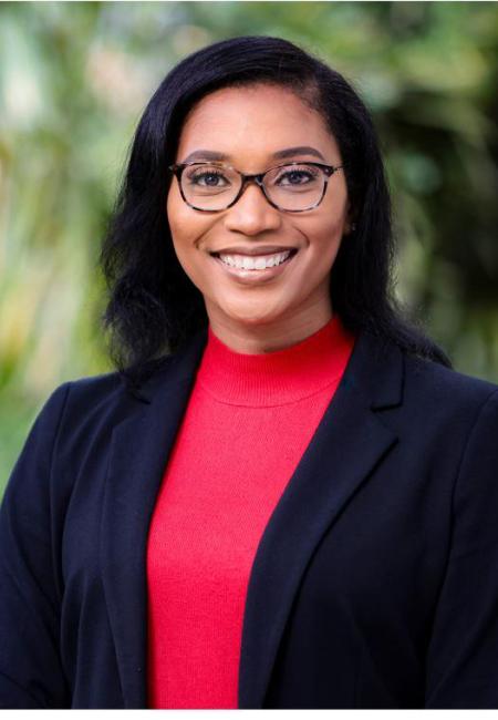 Black woman wearing glasses, a red shirt and a black blazer in front of a green background