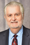 Man in front of grey background wearing dark suit, blue shirt and red tie