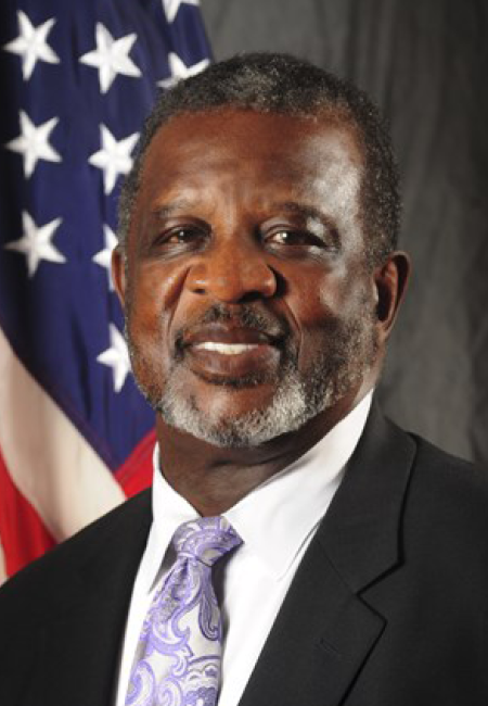 Man in front of US flag in black suite with white shirt and pastel tie