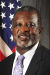 Man in front of US flag in black suite with white shirt and pastel tie