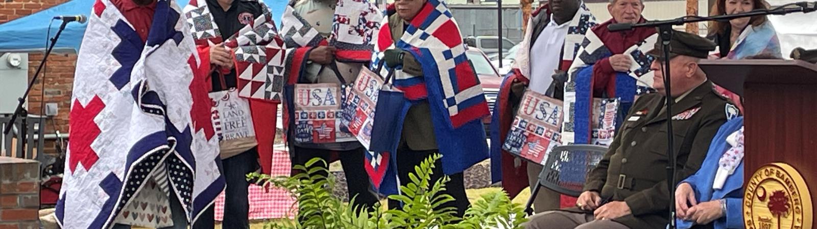 Military veterans stand on a stage, draped in red-white-and-blue quilts made as part of the Quilts for Valor project. 