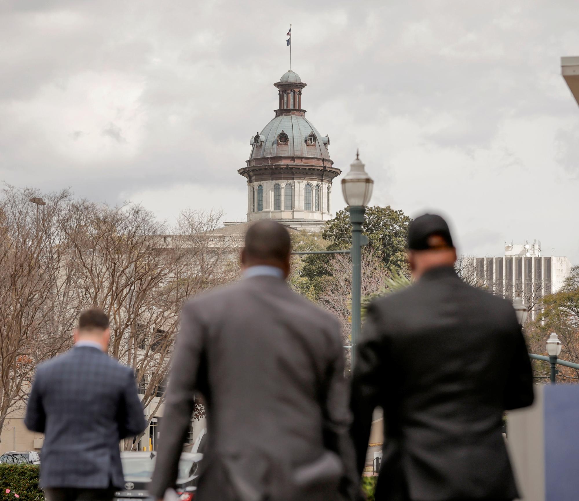Three men in business attire walk toward the SC State House on a cloudy day.