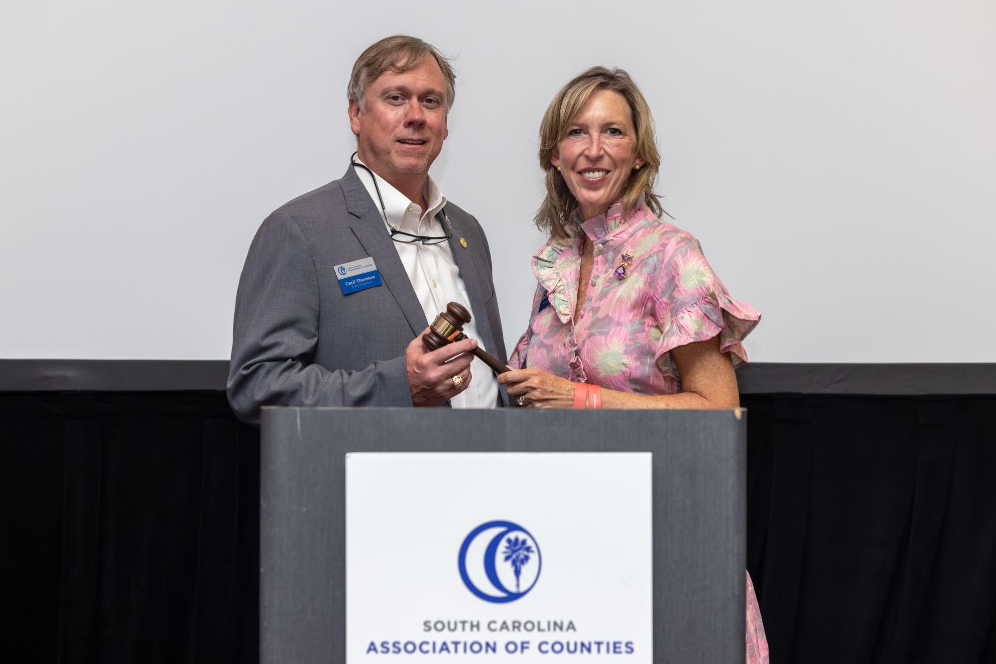 Cecil Thornton and Mary Anderson stand side-by-side holding a gavel behind a podium with the SCAC logo on it.