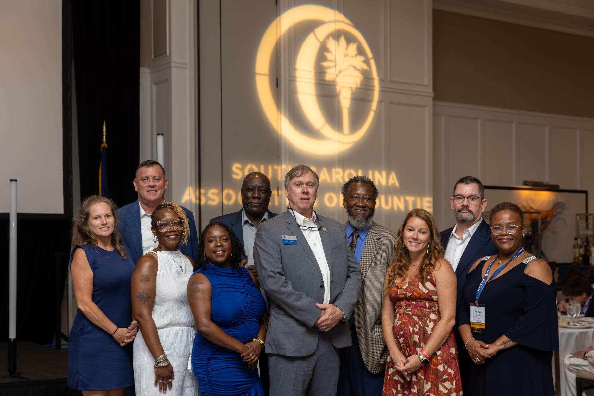 A group of county officials stand in front of the SCAC logo which is projected on the ballroom wall behind them. 