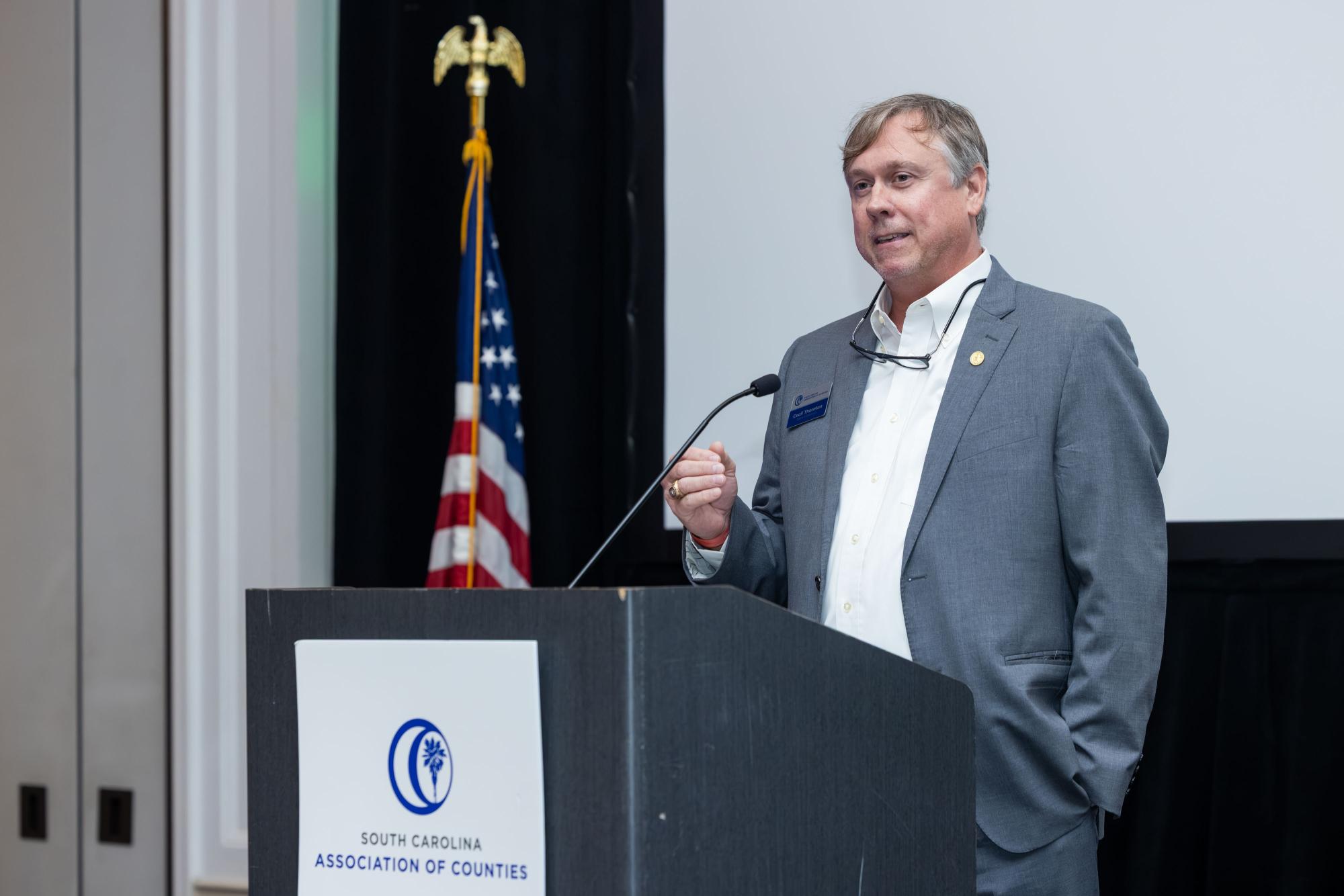 Cecil Thornton stands behind a podium with a microphone. The American flag is in the background.