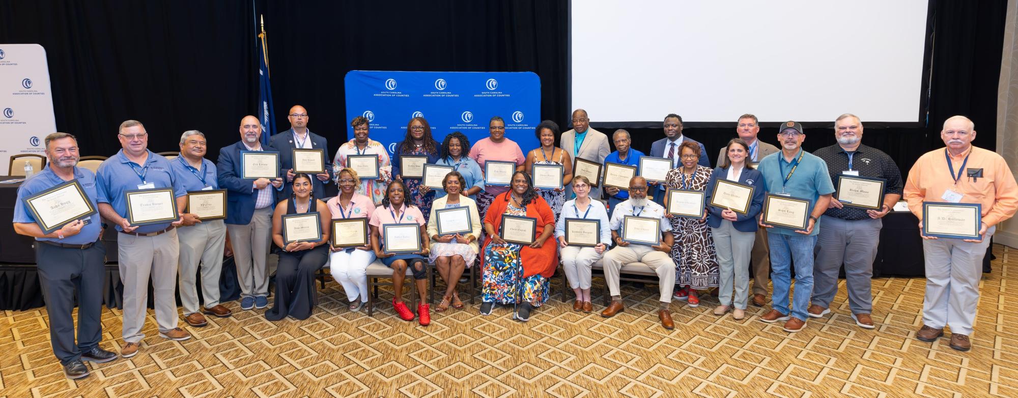 County officials, holding certificates, stand in a group in a ballroom.