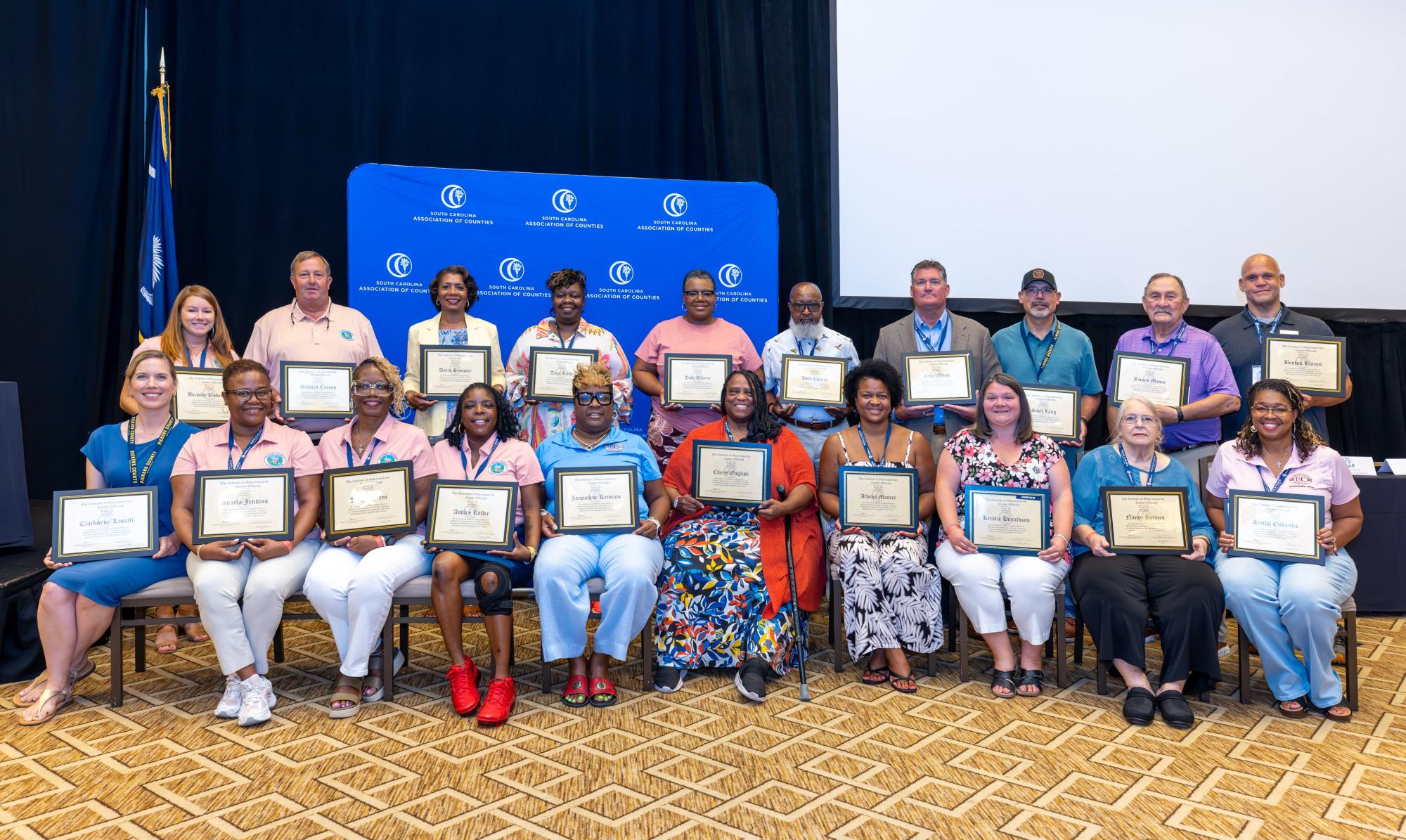 County officials, graduates of the Institute of Government Level 11, stand in a group holding certificates in a ballroom. 