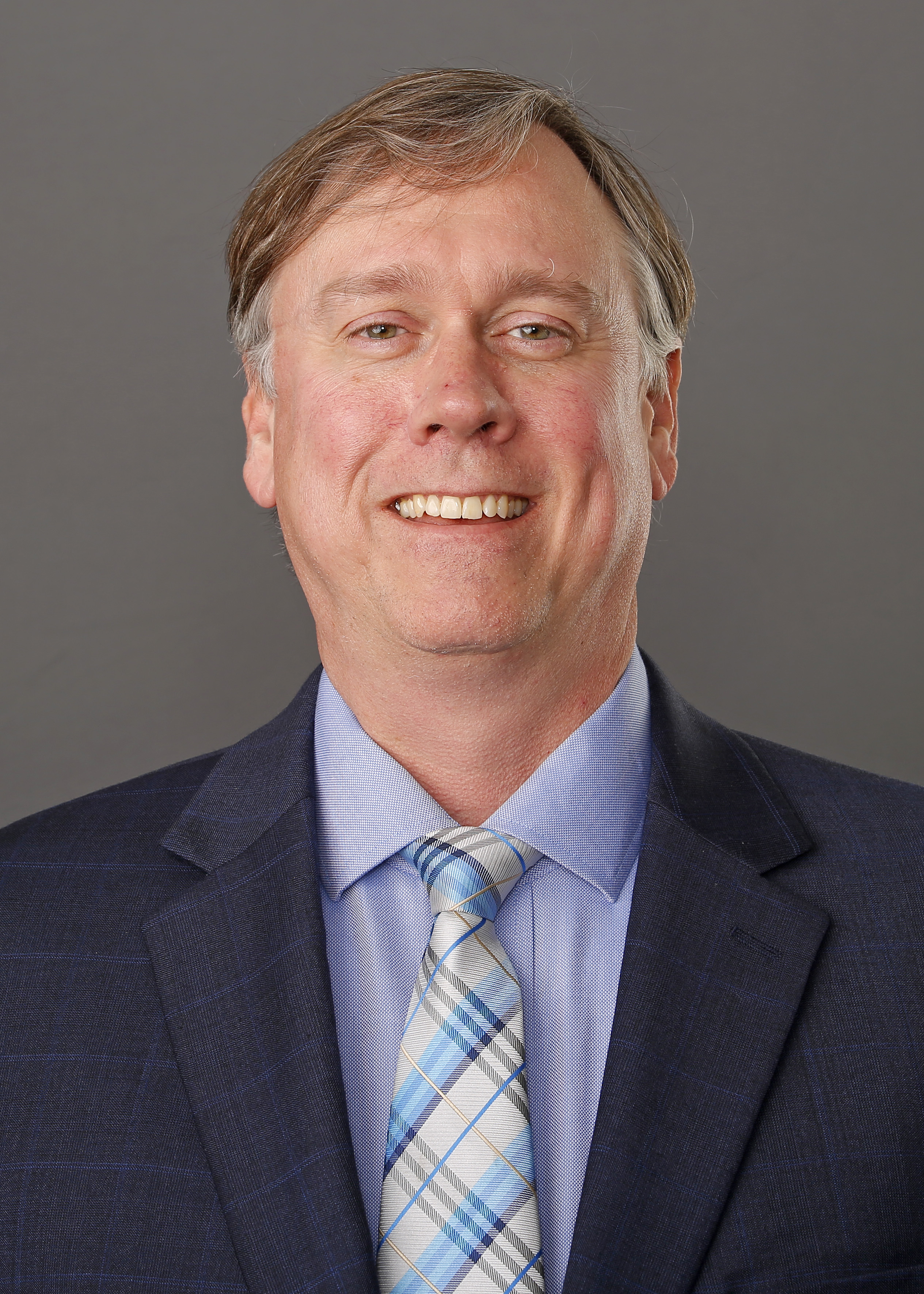 A middle-aged man with light skin and brown hair is smiling. He is wearing a dark suit jacket, a light shirt and plaid tie.