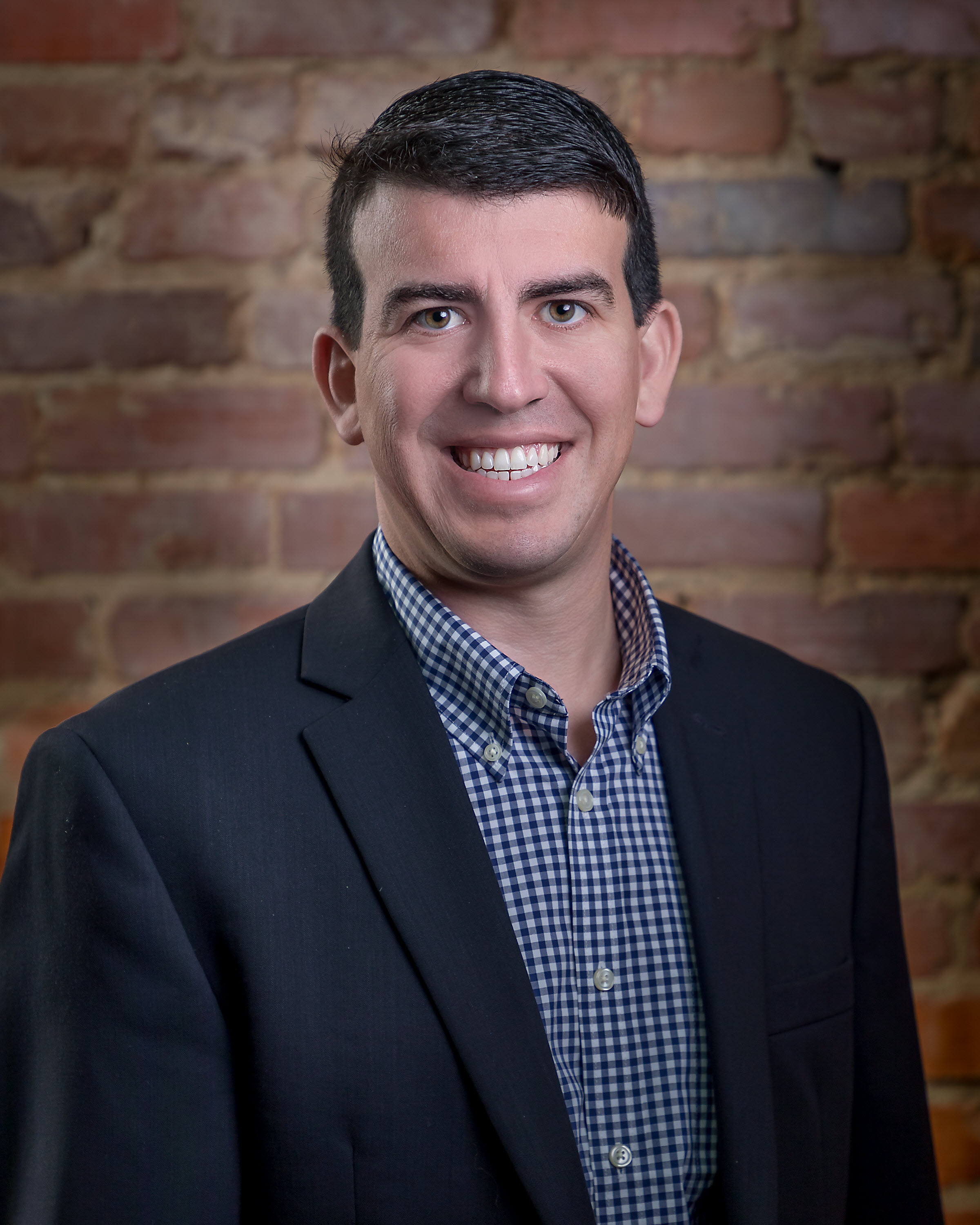 Man in a blue suit jacket with a blue button down shirt in front of a brick background
