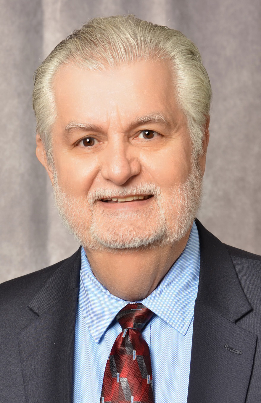 Man in front of grey background wearing dark suit, blue shirt and red tie