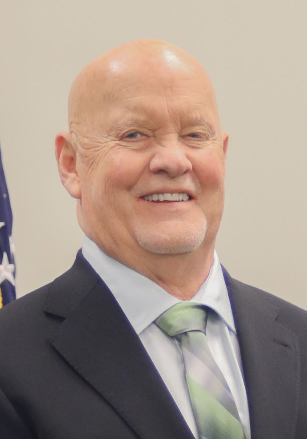Bald white man in dark suit with blue shirt and tie in front of light background