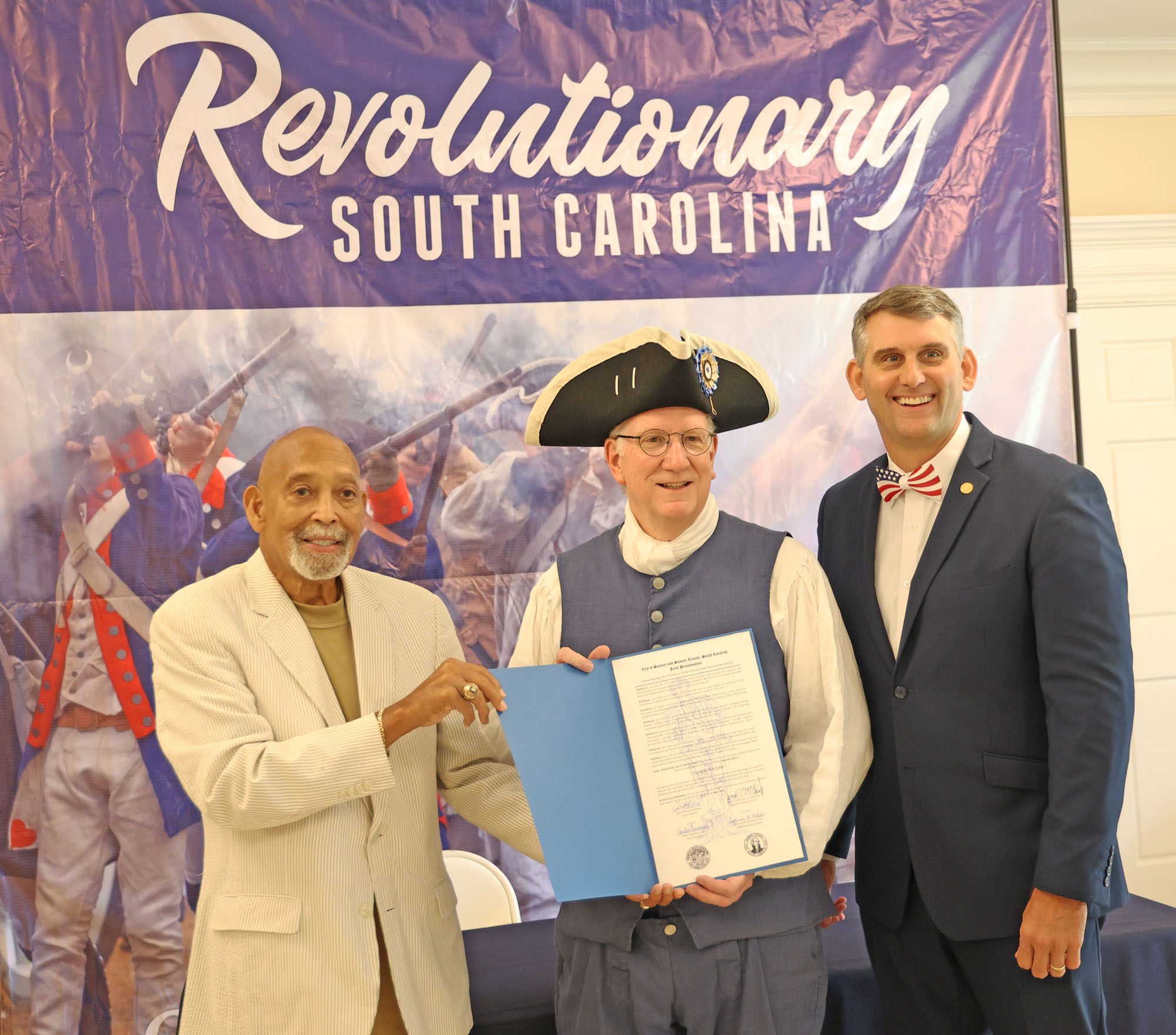 Three men, one in Revolutionary era period costume, stand in front of a banner that says "Revolutionary South Carolina."